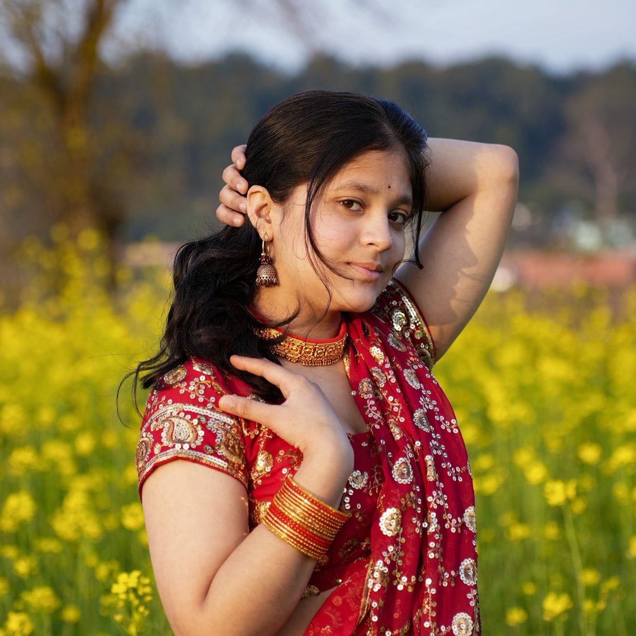A young South Asian woman poses gracefully in a red saree amidst a vibrant mustard field.