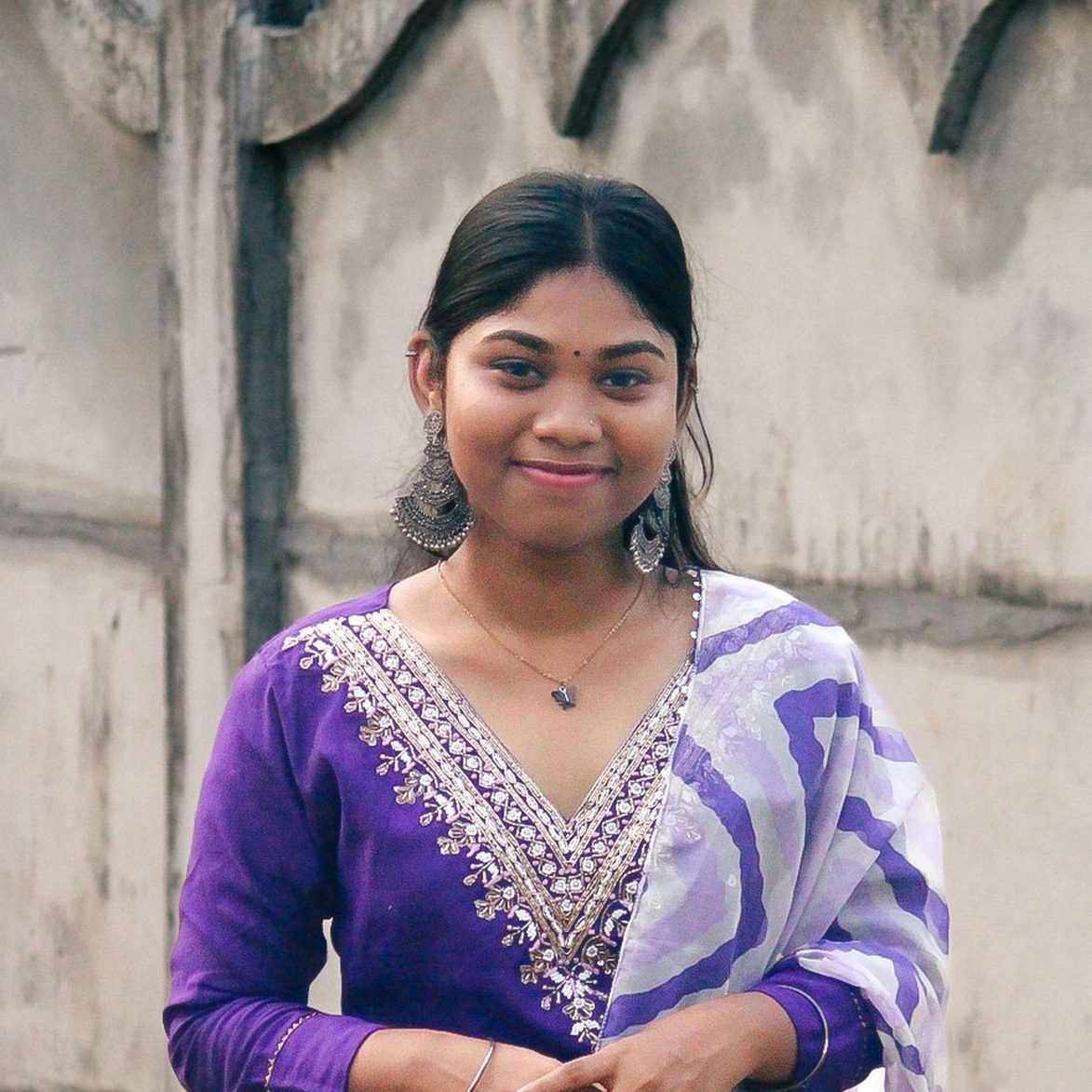 A young South Asian woman smiles outdoors in a vibrant purple saree, embracing cultural fashion.