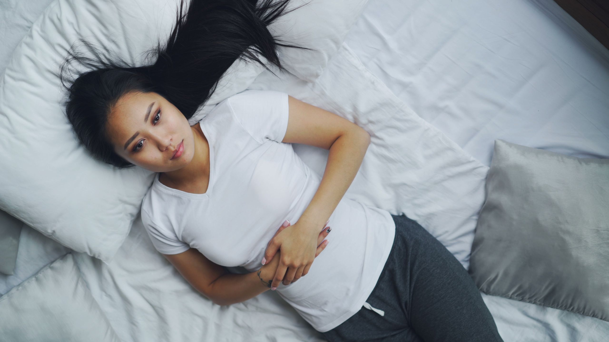 A young woman in casual attire lies on a white bed, looking relaxed and thoughtful.