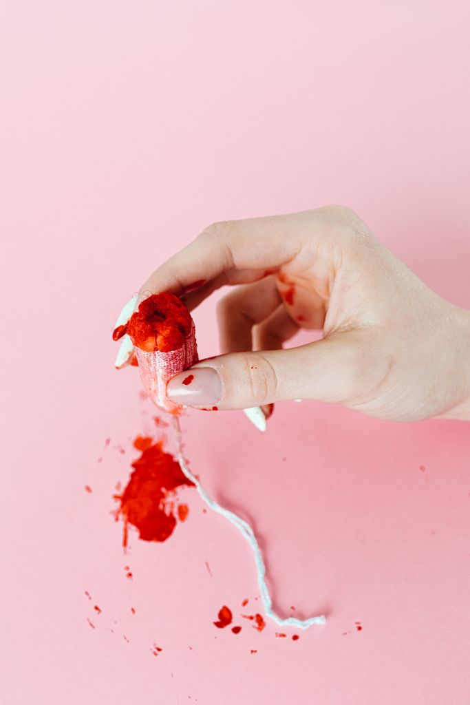 Close-up of a hand holding a used tampon against a pastel pink background, highlighting menstrual themes.